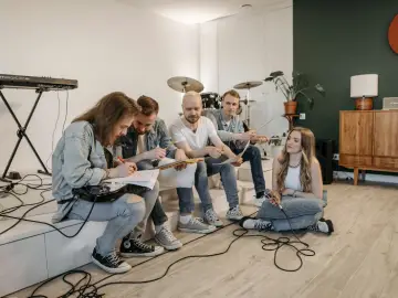 A small group of musicians sitting together in a bright room, discussing song ideas and practising with guitar, microphone, and drums in the background.