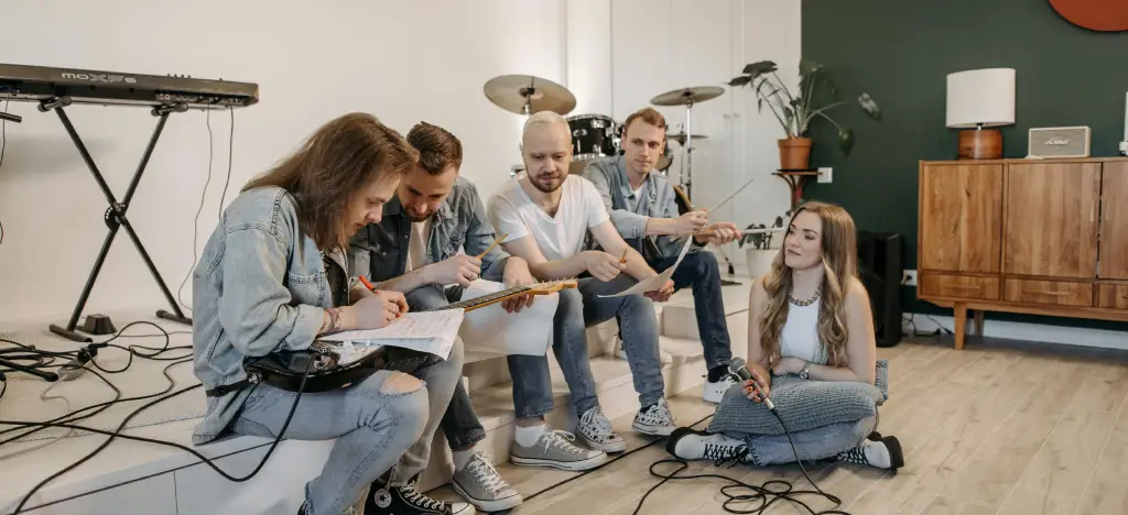 A small group of musicians sitting together in a bright room, discussing song ideas and practising with guitar, microphone, and drums in the background.