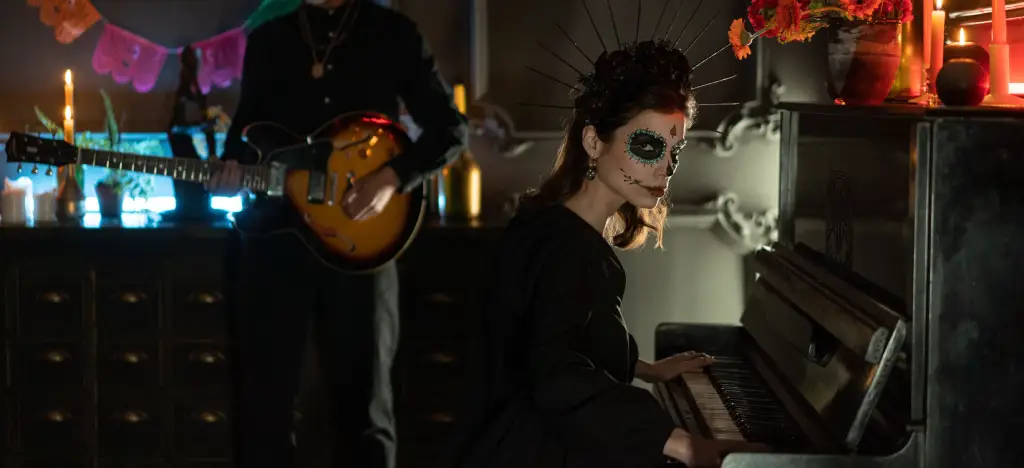 Woman with Day of the Dead skull makeup playing an upright piano at a Halloween party, with a guitarist in the background.