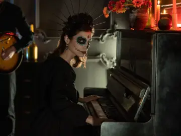 Woman with Day of the Dead skull makeup playing an upright piano at a Halloween party, with a guitarist in the background.